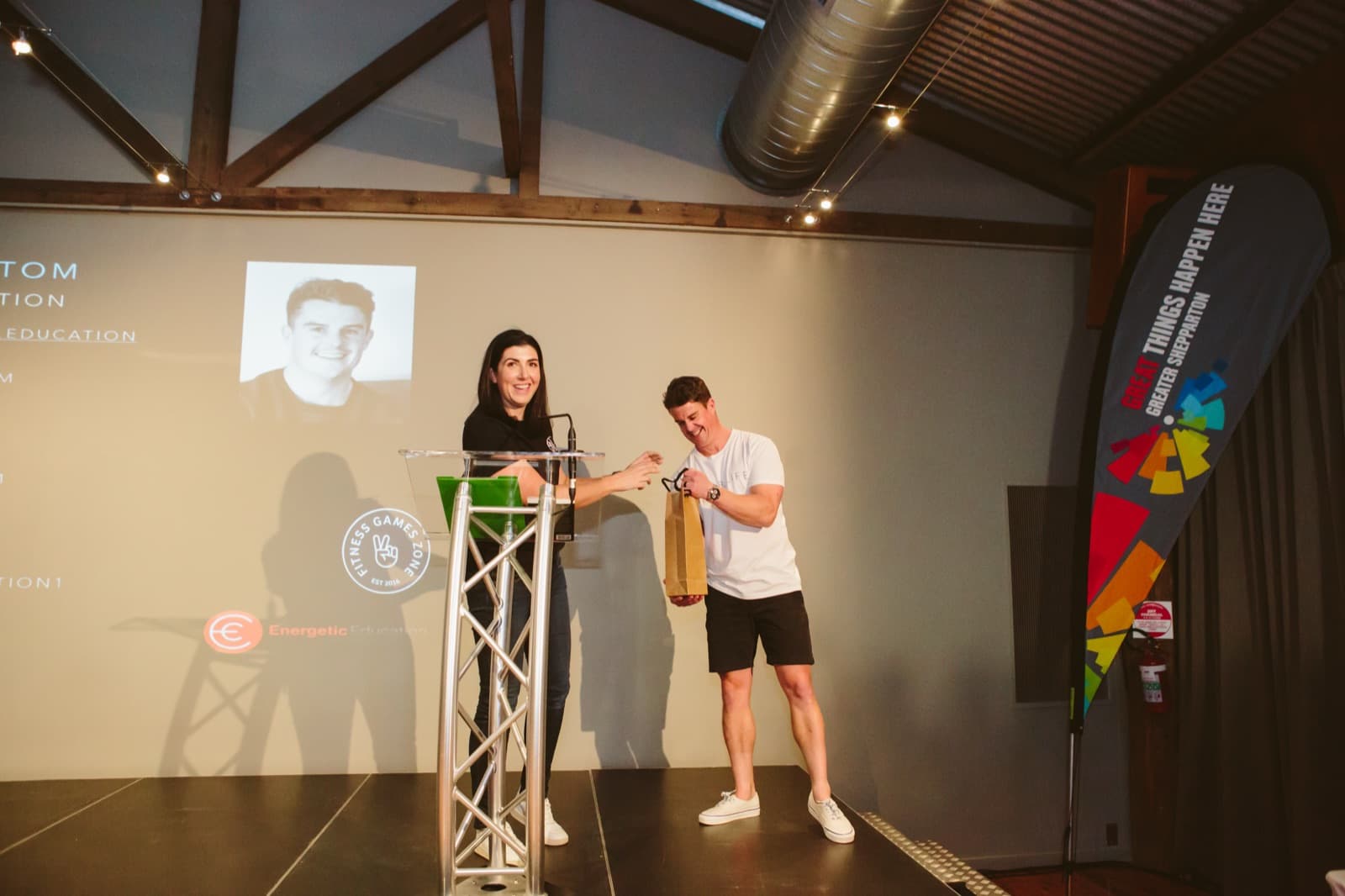 A Better Us Project facilitator on stage beside a host at a lectern, in a timber-beamed venue