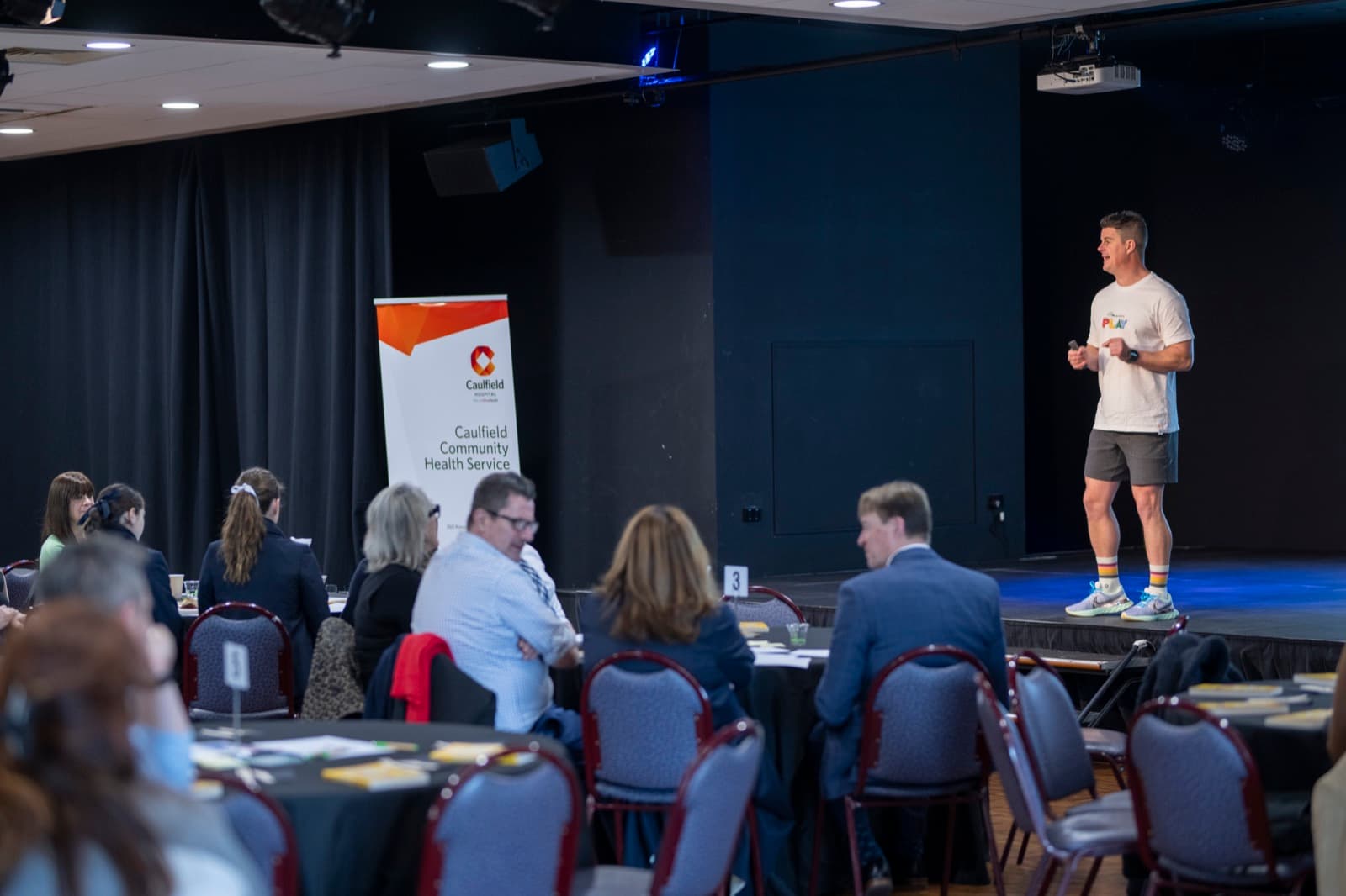 A Better Us Project facilitator on a low stage talking to a team seated at round tables