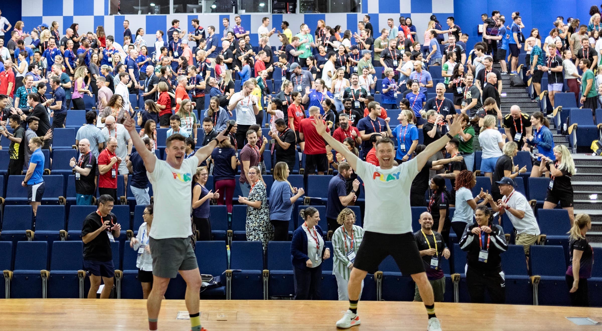 Two Better Us Project facilitators on stage with their arms raised, in front of a full room of people applauding