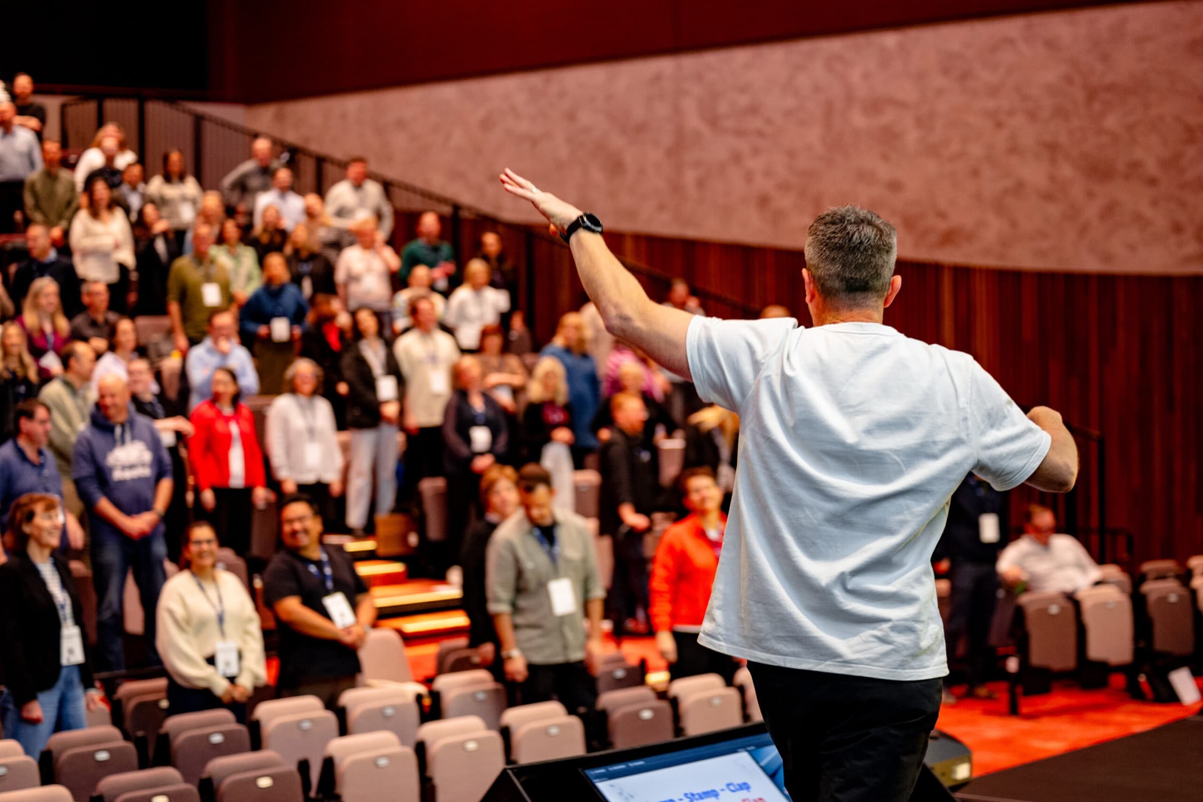 A Better Us Project facilitator on stage with an arm raised, seen from behind, facing a full auditorium of people standing and taking part