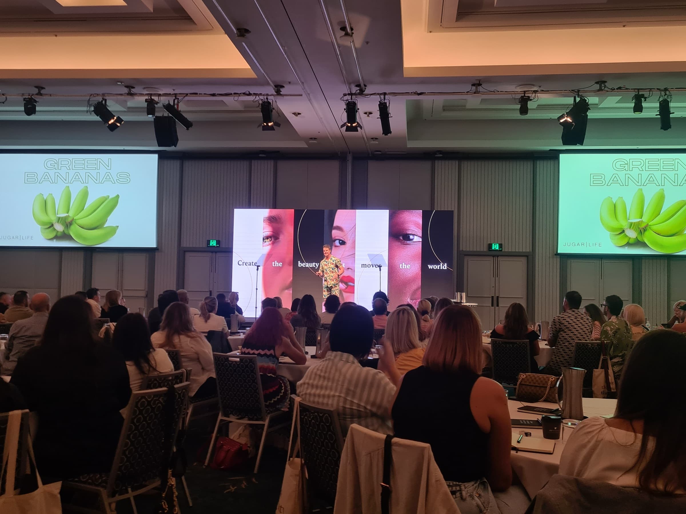 A packed conference ballroom seen from the back, with a speaker on stage between large screens