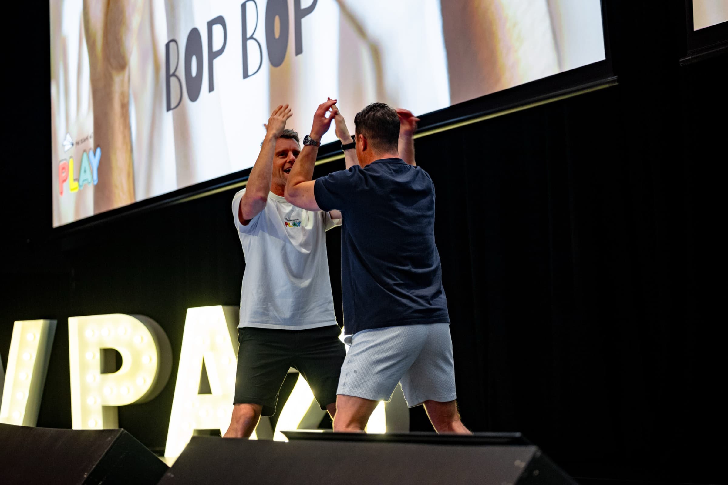 Two Better Us Project facilitators meeting in a high five on stage, in front of a large screen