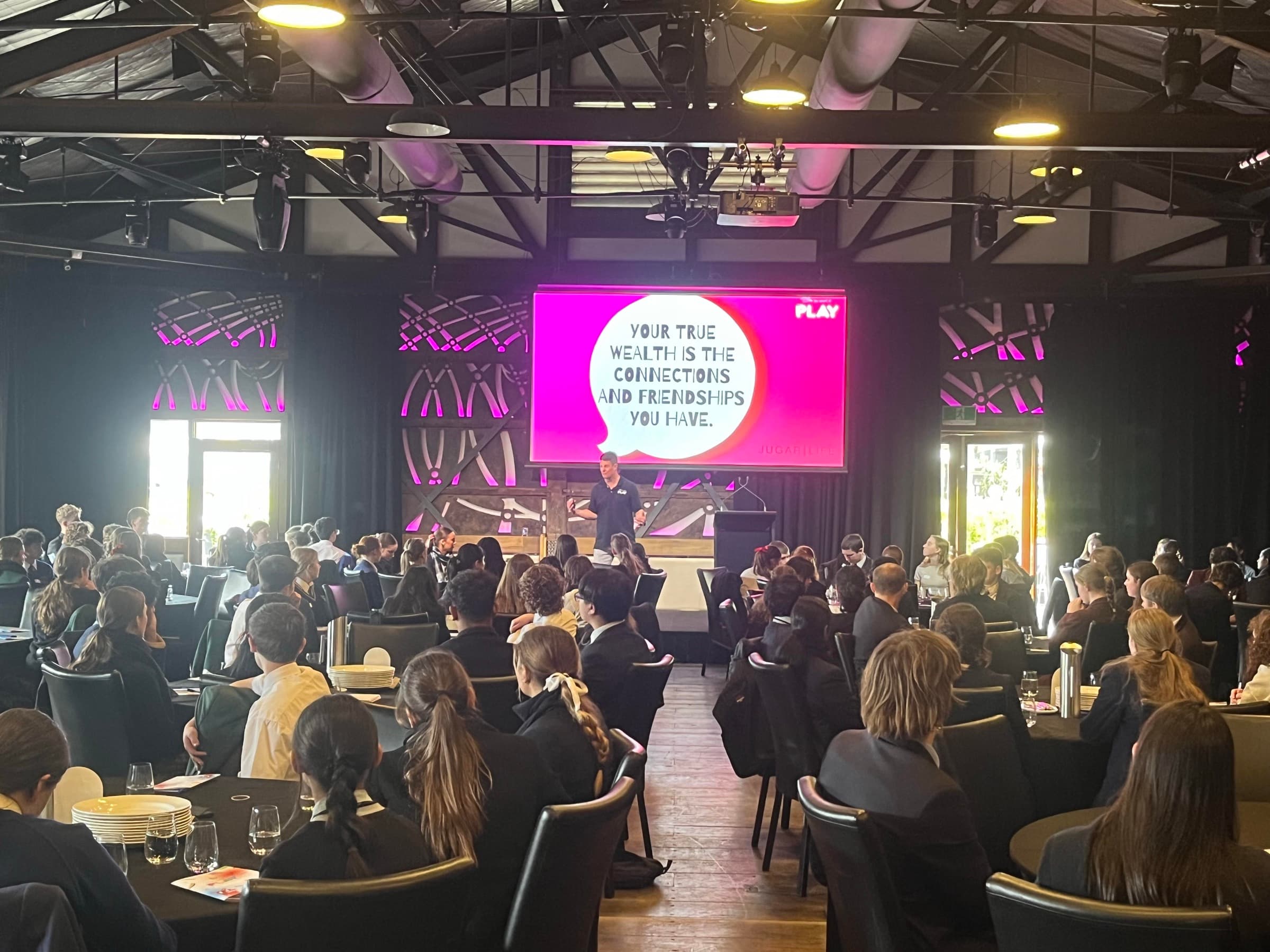 A Better Us Project facilitator speaking to a seated audience at round tables, with a screen behind reading: your true wealth is the connections and friendships you have