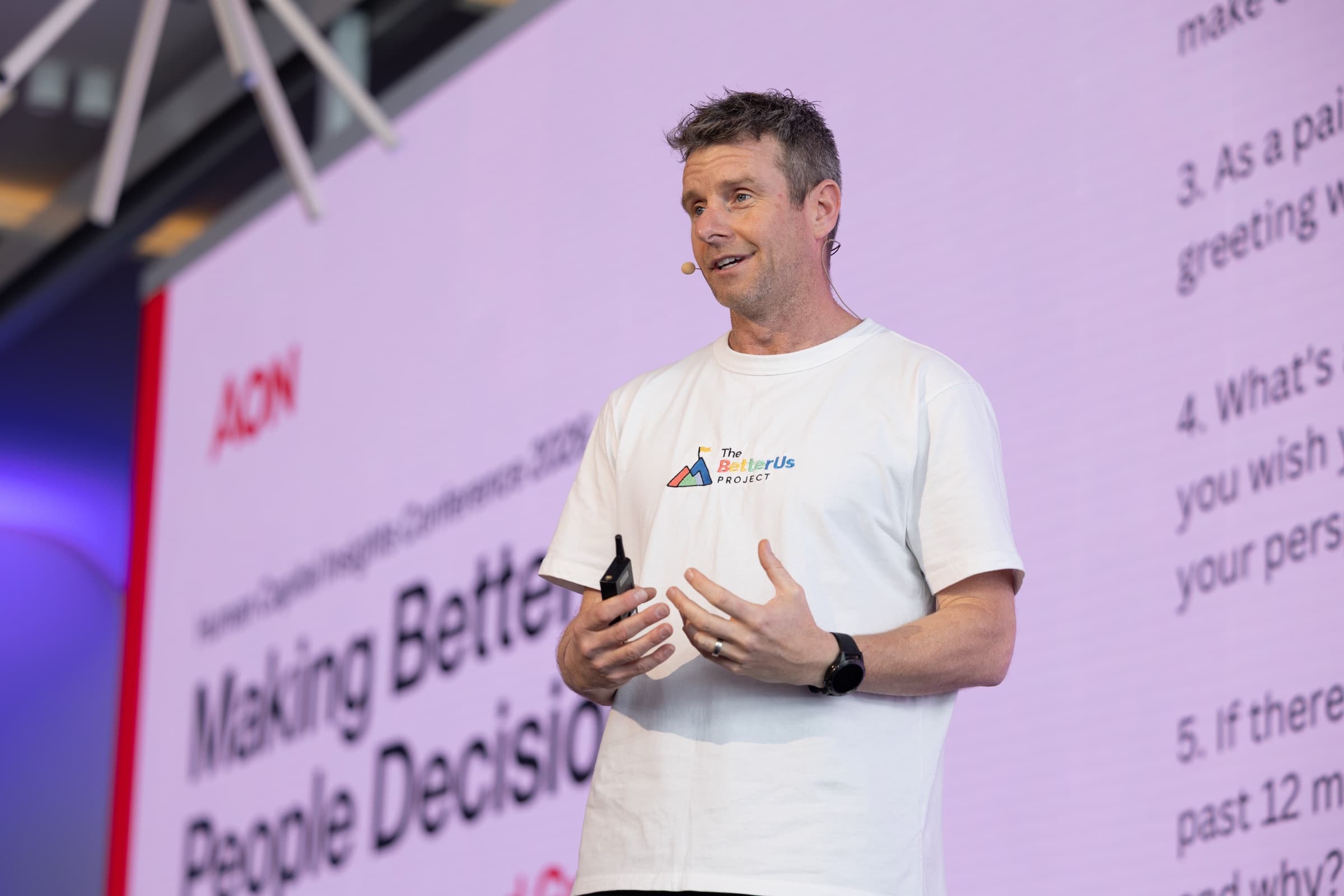 A Better Us Project facilitator speaking on stage in a Better Us Project t-shirt, in front of a large screen at a corporate conference
