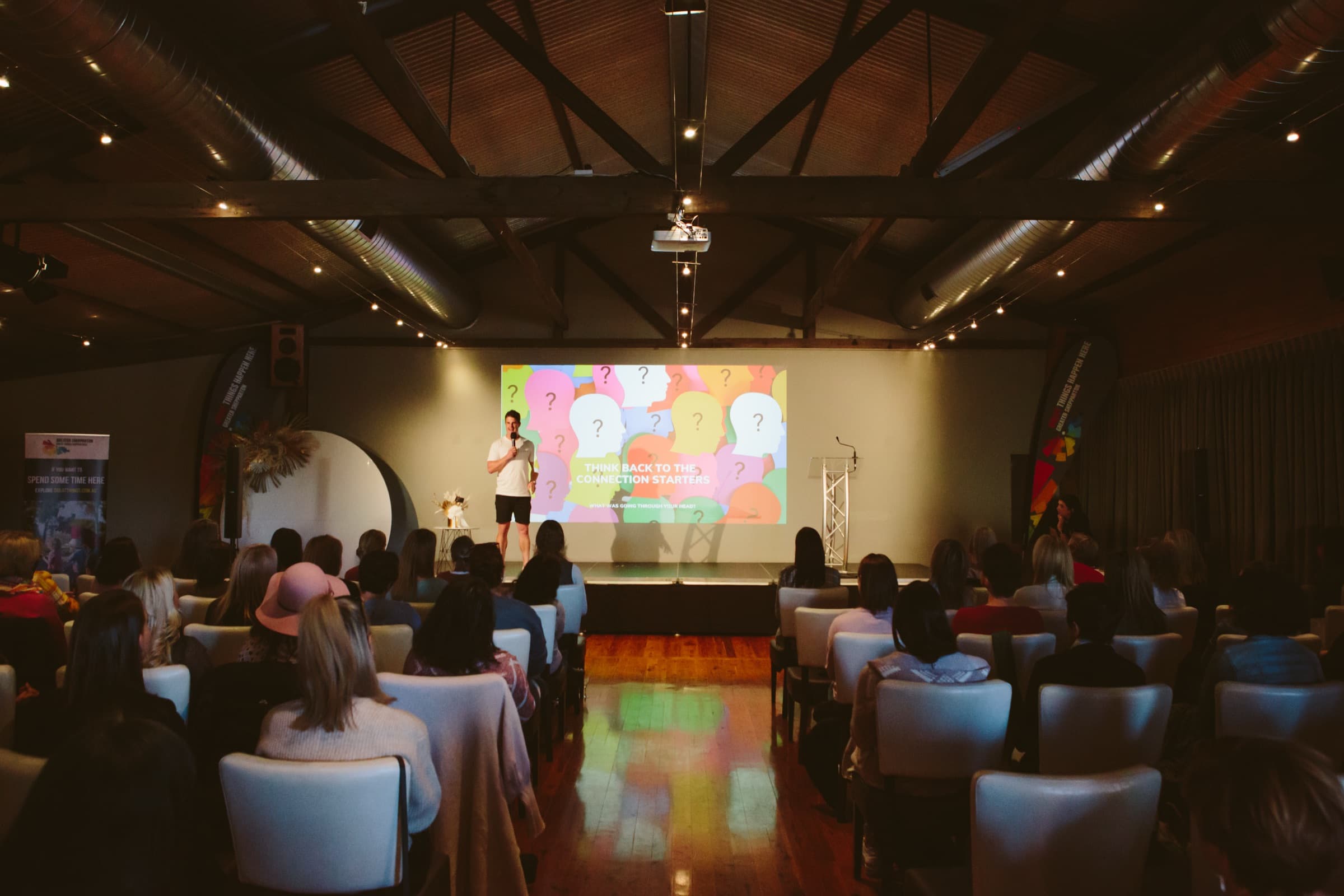 A warm timber-beamed venue with an audience seated in armchairs, watching a facilitator on stage beside a screen