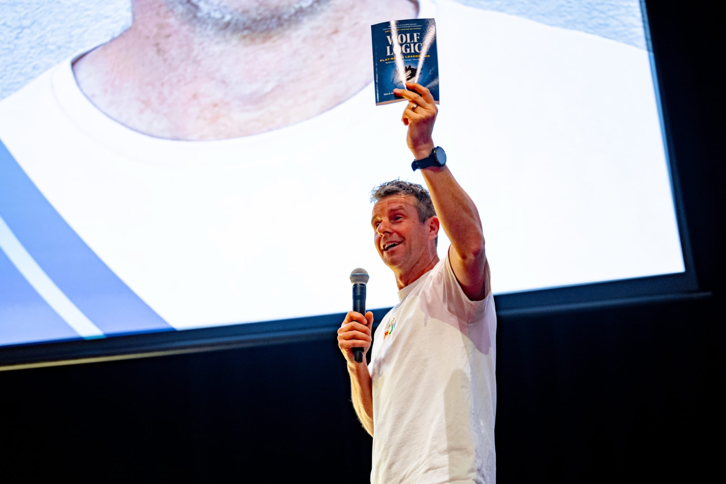 A Better Us Project facilitator on stage holding a copy of Wolf Logic above their head while speaking into a microphone